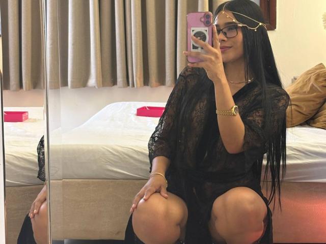 AntonellaPreschot with long black hair, glasses, and gold chain, squatting in front of mirror, wearing sheer black dress, holding pink phone, showing knees, and smiling.