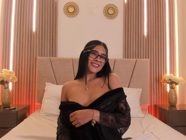 AntonellaPreschot with black hair, glasses, and a smile, sits on a bed, pulling down a black lace top to reveal her tits, in a warmly lit bedroom.