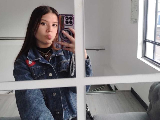Melani Becker mit langen, braunen Haaren, voller Lippen, trägt eine Jeansjacke, ein schwarzes Hemd und eine Halskette und macht ein Selfie im Spiegel, wobei sie ein lila Handy mit einem Ringgriff hält..