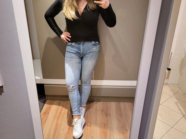Linnea-hot with blonde hair, black long-sleeve top, and ripped blue jeans, standing in a mirror, showing her ass and white sneakers, holding a phone.