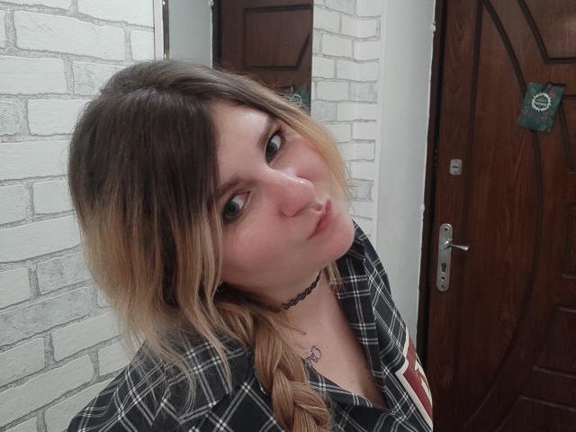 MinistraX with blonde and brown ombre hair, plaid shirt, black choker, pouty expression, and small heart tattoo on chest, standing in front of a wooden door.