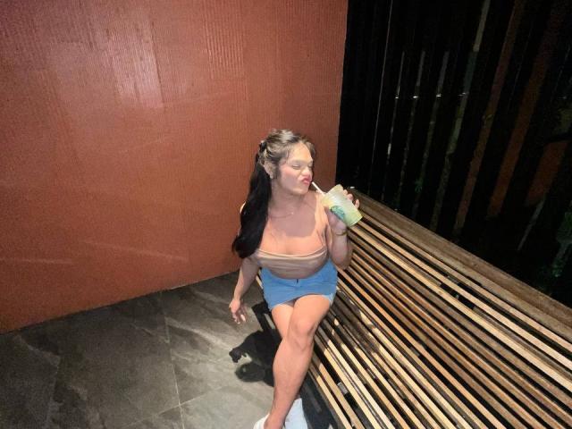 EsaMarie, a young woman with fair skin and long black-and-brown hair, sits on a wooden slatted bench. She's wearing a beige strapless top and blue shorts. She holds a green drink in her right hand and looks to the left. The wall behind her is red, and the floor is dark gray. Her left leg is bent at the knee, and she's wearing white sneakers. The image is a photograph.