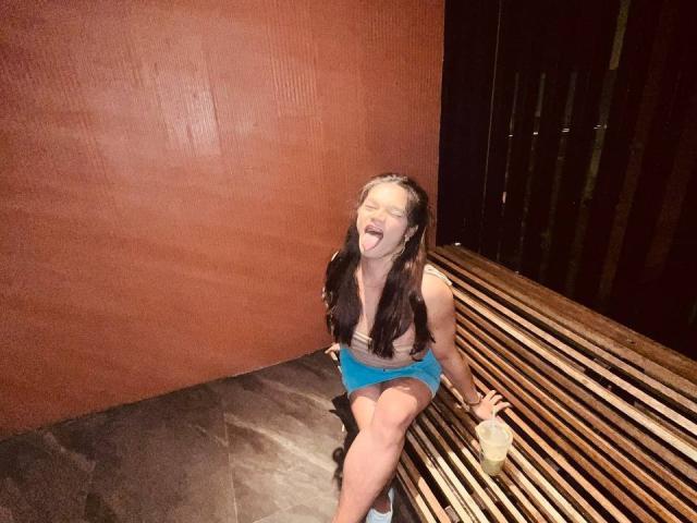 EsaMarie, with long dark hair, tongue out, eyes closed, wearing a blue top and shorts, sitting on a wooden bench, holding a drink, in a dimly lit room.