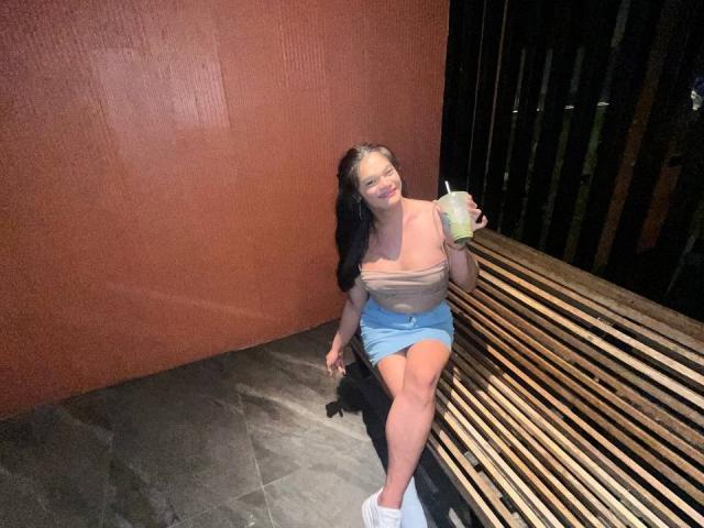 EsaMarie with long black hair sits on a wooden bench against a red textured wall. She's wearing a beige off-shoulder top and blue mini skirt, holding a green drink with a straw in her right hand. She has white sneakers on. EsaMarie's smile is visible, and she's looking slightly to the left. The bench has slats, and the floor is gray tile. The lighting is even, highlighting her.