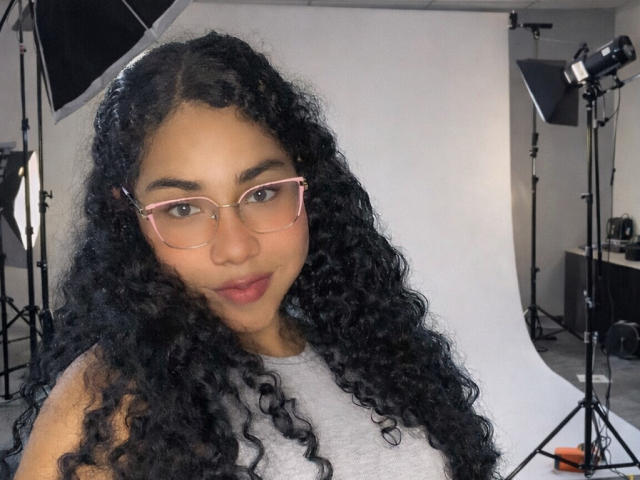 EmaFfox with curly black hair, light brown skin, pink glasses, gray sleeveless top, and a slight smile, standing in a studio with photography lights in the background.