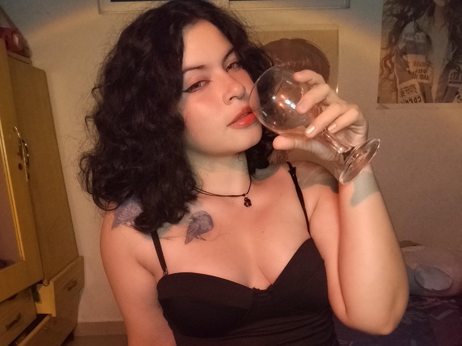 SarahLawrens with curly black hair, black strapless top, holding a wine glass, pink nose ring, black choker, small tattoo on right shoulder, pale skin, red lips, slightly open mouth.