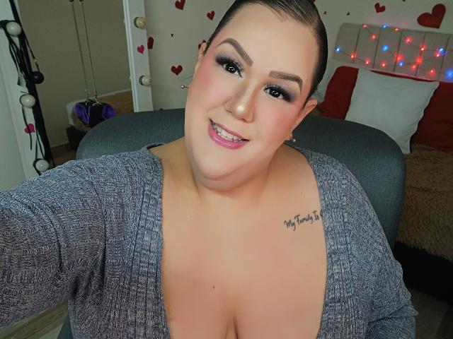 GiiannaMorrys, avec de grandes seins, portant un pull gris, souriant à la caméra, a un tatouage noir sur la poitrine droite, des cheveux bruns attachés en chignon, du rouge à lèvres rose et un maquillage des yeux prononcé..
