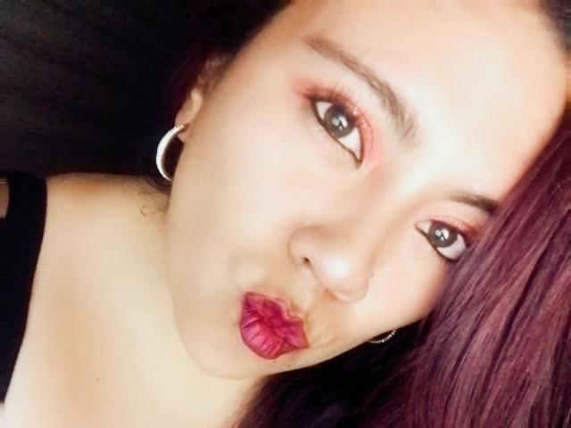 Close-up photo of LexyDom with medium skin tone, dark brown hair, and subtle pinkish-red eyeshadow. She has dark, arched eyebrows, small silver hoop earrings, and wears bold pink lipstick. LexyDom's eyes are slightly open, and her lips are puckered in a playful, kissing gesture. She's wearing a black strap, visible on her shoulder. The composition is slightly tilted, focusing on her face. The lighting is soft, highlighting her facial features. Her expression is playful and engaging.