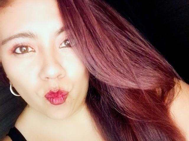 Close-up selfie of LexyDom with light brown skin, long red-brown hair covering one eye, and dark brown eyes. She's puckering her lips with bright red lipstick. LexyDom wears small silver hoop earrings. The background is dark, making her face and hair stand out. The light highlights her smooth skin and hair texture. The image has a casual, candid feel.