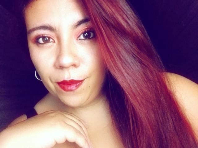 Close-up of LexyDom with medium brown skin and long, straight red hair. LexyDom's brown eyes are slightly upturned, and she has dark, well-groomed eyebrows. She wears red lipstick and has a small nose piercing on her left nostril. Her right hand is resting near her chin. She has hoop earrings. The lighting is even, highlighting her facial features. The background is dark and indistinct. The image is slightly overexposed, giving a soft, almost glowing effect to her skin.