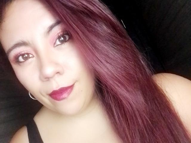 Close-up selfie of LexyDom with light skin and long, straight, dark red hair. LexyDom has pinkish eye shadow, dark eyeliner, and bright red lipstick. She's wearing a black strap top and small silver hoop earrings. LexyDom's face is slightly angled to the right, with a subtle, confident smile. The light source is from the front, highlighting her facial features. The background is dark and blurred.