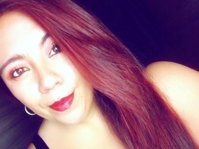 Close-up of LexyDom with long, straight, reddish-brown hair covering the right side of her face. She has fair skin, dark eyebrows, and brown eyes with pink eyeshadow. LexyDom wears red lipstick and a black sleeveless top. She has a small nose piercing and a silver hoop earring in her left ear. The background is dark, making LexyDom's face and hair stand out. The image has a slightly overexposed, bright quality. LexyDom smiles subtly, looking at the camera.