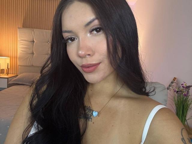 BerlyVenuss has long, dark, wavy hair and a fair skin tone. She wears a white strap top and a blue heart necklace. Her brown eyes are slightly almond-shaped, and she has a subtle, confident smile. She has a small tattoo on her right shoulder and a nose ring. The soft light highlights her smooth skin and relaxed posture. The background includes a bed with a tufted headboard and a lamp on a nightstand.