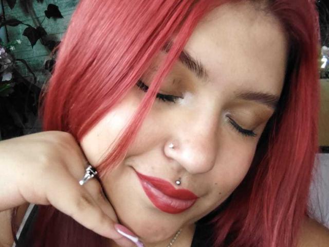 AmberCutel with red hair, nose piercings, and red lipstick, eyes closed, smiling, hand touching chin, wearing a ring with a small diamond, and a nose stud.