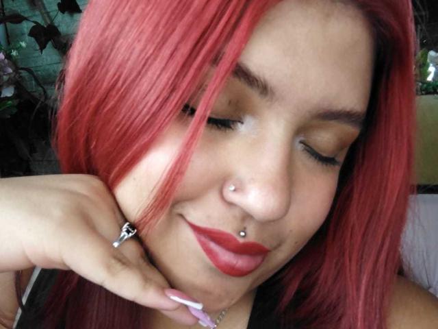 AmberCutel with red hair, nose piercing, and red lipstick, eyes closed, smiling, holding her chin, showing a silver ring, and pink nails, with a nose ring.