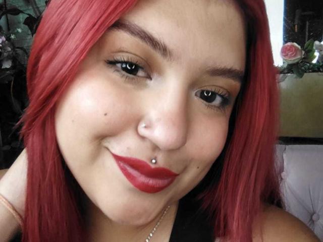 AmberCutel has red hair, a nose piercing, red lipstick, and a small black mole on her left cheek. She's smiling with black eyeliner and a silver necklace.