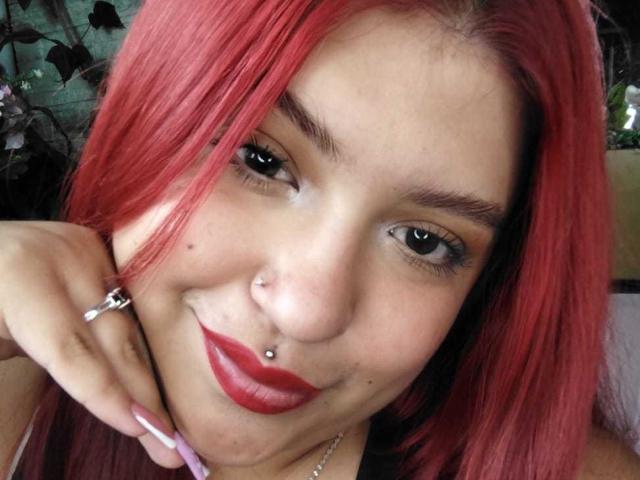 AmberCutel with red hair, nose piercings, and red lipstick, smiling, showing a septum piercing, and holding her chin with purple nails. Her tits aren't visible.