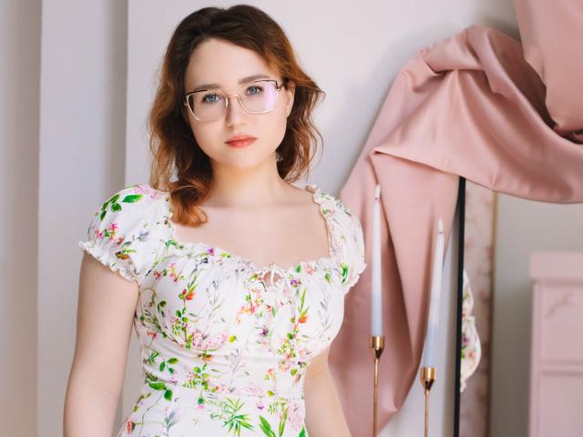 TeresaBirt with wavy brown hair, glasses, white floral dress, small tits, pink jacket on a stand, light skin, standing, looking at camera, soft pink background.