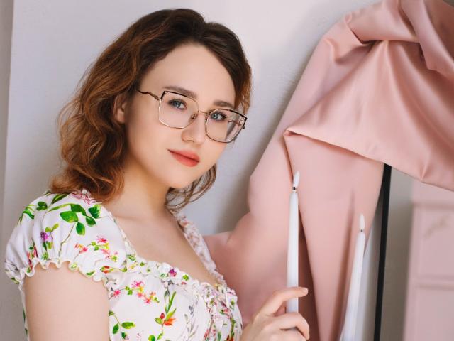 TeresaBirt, a young woman with fair skin and wavy, brown hair, wears glasses with thin, black frames. She has a subtle, pinkish lipstick and a slight, confident smile. She holds a pink jacket on a white hanger with her right hand. She wears a white dress with green and pink floral patterns, with ruffled sleeves. The dress has a slightly loose, comfortable fit. The jacket is a soft, pastel pink with a smooth texture. The composition is centered on TeresaBirt, with the jacket to her right and the hanger slightly out of focus. The overall style is casual and natural.