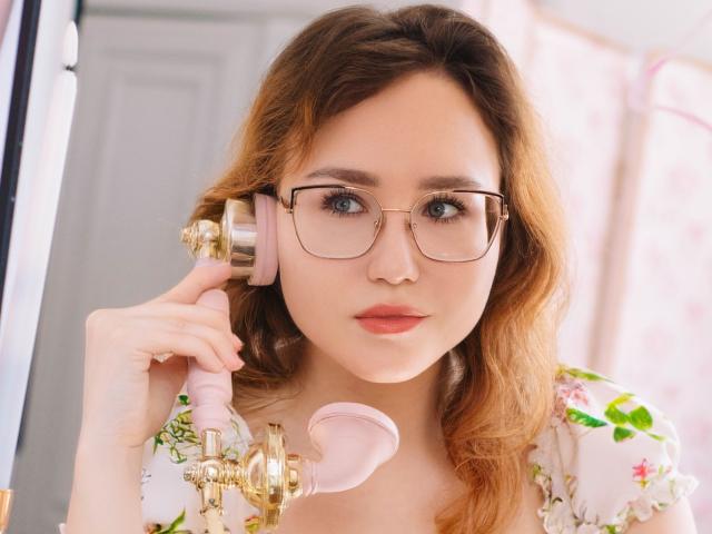 TeresaBirt with wavy brown hair, glasses, pink hairphone, and floral top, holding a pink phone, looking directly at the camera with a neutral expression.