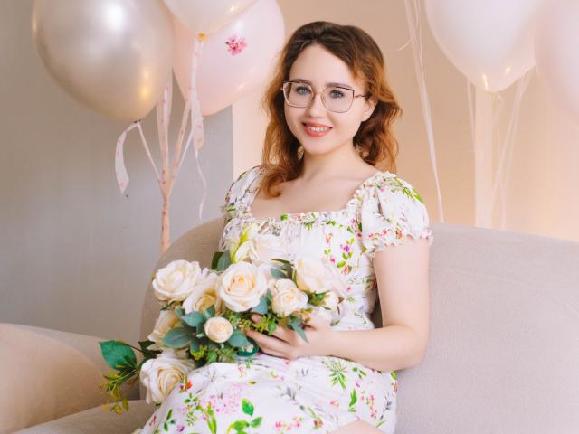 TeresaBirt sits on a beige sofa, holding a bouquet of white and yellow roses. She has fair skin, wavy light brown hair, and wears glasses. Her floral dress has white with pink and green patterns. The background includes white and silver balloons with pink ribbons. The soft lighting highlights her smile and the bouquet's delicate petals. Her relaxed posture contrasts with the delicate, elegant composition. The image uses a natural, candid style.