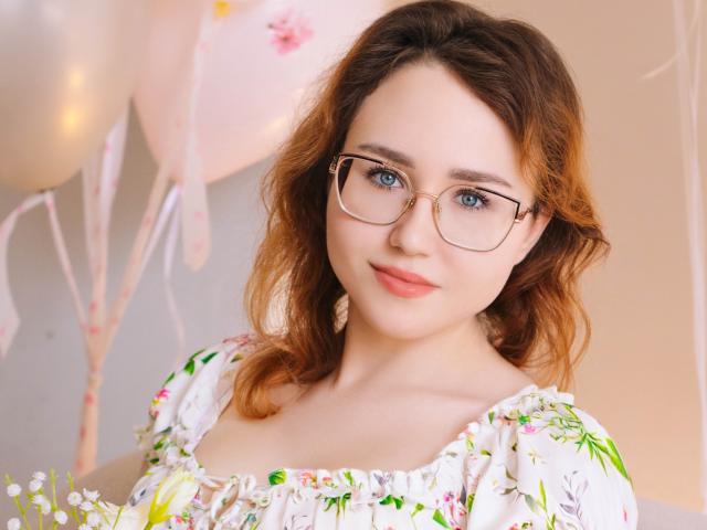 Portrait of TeresaBirt with fair skin and wavy, medium-brown hair. She wears thin, gold-rimmed glasses and a floral, white dress with green and pink patterns. Her blue eyes are slightly open, and she has a soft, pink lipstick. The background has blurred, pastel balloons and soft lighting. The composition focuses on her face and upper chest. The image has a warm, natural light. The floral dress and balloons add a festive, gentle atmosphere.