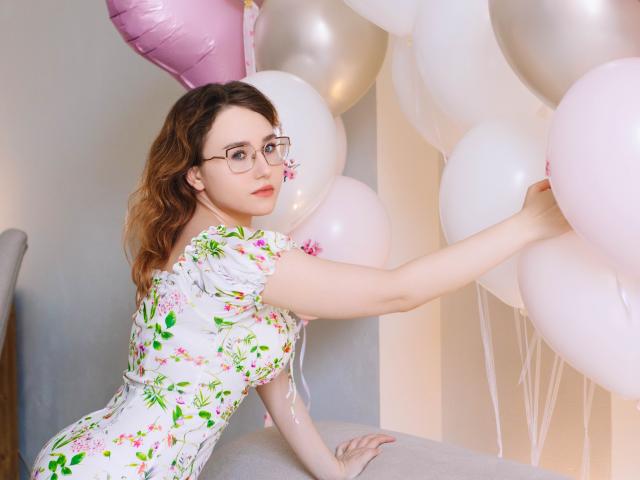 TeresaBirt with wavy brown hair and glasses, wearing a white floral dress, reaches into a cluster of pink and white balloons. Her right arm extends, gripping balloons on the right. Soft lighting highlights her fair skin and floral dress. The balloons have a shiny, reflective texture, contrasting with her matte dress. Her expression is calm, with slightly parted lips. The image has a balanced composition with TeresaBirt on the left.