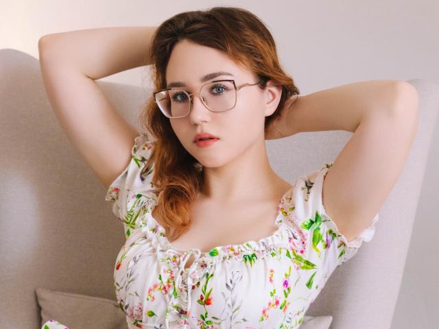 TeresaBirt, a young woman with fair skin and wavy auburn hair, wears clear-framed glasses and a floral, off-the-shoulder, white dress with puffed sleeves. She has her arms raised, hands behind her head, and gazes directly at the camera with a slightly parted, pink-lipped mouth. Her dress features green, pink, and white floral patterns. The light gray armchair she's seated in is slightly out of focus. The composition is centered, emphasizing TeresaBirt's relaxed yet confident posture. Her soft, natural makeup enhances her delicate features. The overall image is bright and clear, highlighting her youthful, attractive appearance.