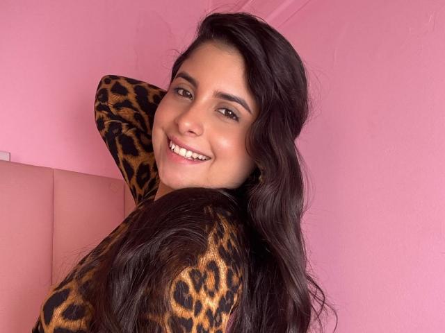 Luciana Blake, com cabelos longos e escuros, sorrindo para a câmera, vestindo uma blusa com estampa de leopardo, exibindo seus seios, em frente a uma parede rosa. Seu braço está levantado atrás da cabeça..