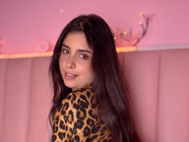LucianaBlake with long dark hair, smiling at the camera, wearing a leopard print top, looking over her shoulder, pink background, soft lighting, slight blush on her cheeks.