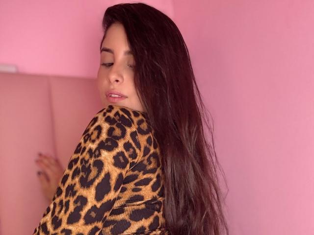 Luciana Blake, con cabello largo y castaño oscuro, vistiendo una blusa de estampado de leopardo, mirando por encima del hombro, con los ojos cerrados, un muro rosa de fondo, una sonrisa sutil, un cojín rosa claro detrás..