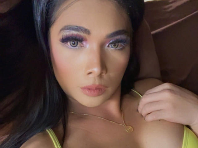 AsianDreamHoney has long black hair, purple eyelids, and pink lipstick, with a gold necklace and green top, lying down, looking at the camera, with a serious expression.