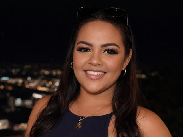 Rizantella has dark brown hair, smoky eye makeup, and a smile showing white teeth. She wears a black top, hoop earrings, and a necklace with a small pendant.