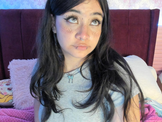 AmaraKazumi is a young woman with long, wavy black hair and pale skin. She has blue eyes, heavy black eyeliner, and pronounced eyelashes. She wears a silver nose ring and has a small, pale pink lip piercing. She's dressed in a gray t-shirt and has a necklace with small blue beads. Her freckles are visible on her cheeks. The background includes a pink fluffy pillow, a yellow pillow with a character design, and a pink bed with a patterned blanket. The headboard is dark red.