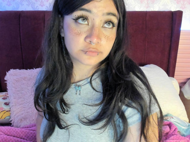 AmaraKazumi has long black hair, a septum piercing, a lower lip piercing, and freckles. She's wearing a grey shirt, looking up with big eyes. Her tits are not visible.
