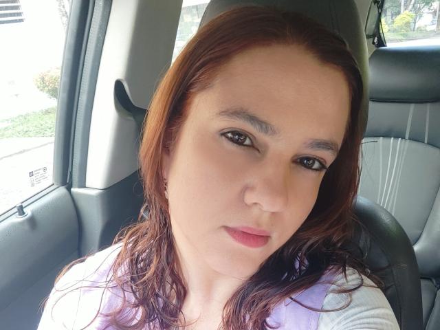 AmeliaWilson has brown hair, light skin, brown eyes, and pink lips. She's wearing a white shirt, sitting in a car, looking at the camera with a neutral expression.