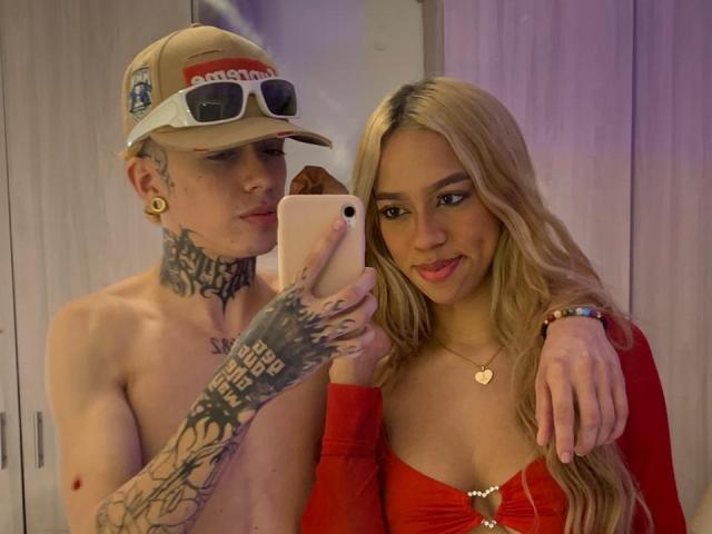 A selfie photograph of IceCumHot and another person. IceCumHot, shirtless, has tattoos covering his neck and hands, wears a beige cap with "adidas" and sunglasses, gold ear studs. He's holding a phone with his tattooed hand. The other IceCumHot has long blonde hair, red off-shoulder top, heart-shaped necklace, and smiles. Both are close, with IceCumHot's arm around the other. The phone is white, and IceCumHot's tattoos are detailed. The image is focused on their upper bodies and faces.