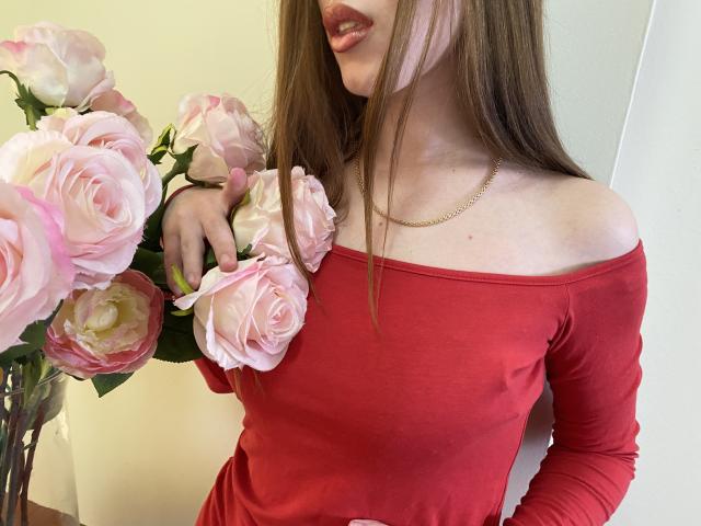 StellaNoir in a red off-shoulder top, holding pink roses, with long brown hair, wearing a gold necklace, showing her tits and a hint of her ass.