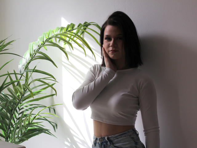 SaraJerome with dark hair, white long-sleeve crop top, and blue jeans, standing against a wall, hand on cheek, green plant to the left, sunlight casting shadows.