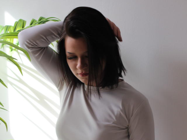 SaraJerome with dark brown bob, white long-sleeve shirt, arm raised, touching head, green plant to left, soft sunlight, slight shadow, serious expression, upper body shot.