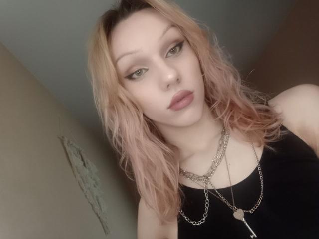 KiranaHayes has shoulder-length, wavy pink hair with darker roots. She has light skin, green eyes with pronounced eyeliner, and full, pink lips. She wears a black sleeveless top and two silver necklaces. The image is a close-up selfie, slightly angled. Her expression is neutral, looking directly at the camera. The background is blurred and indistinct. The lighting is soft, emphasizing her facial features and hair texture.