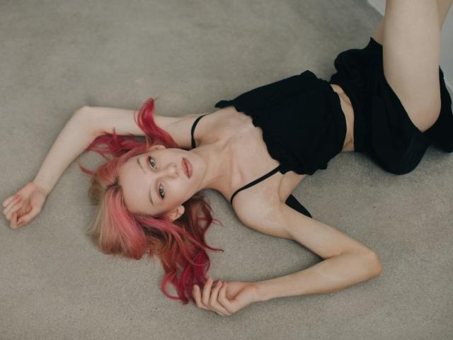 KiranaHayes is lying on a carpet, wearing a black crop top and shorts, with pink and blonde hair, arms and legs spread, showing small tits and a slim, pale body.