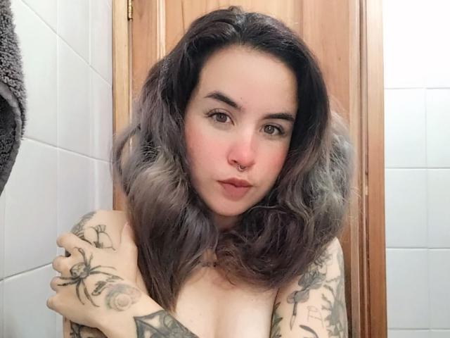 SammyBeil with gray ombre hair, nose ring, and tattoos, covering her bare tits with arms, looking at camera with pouty lips, in a bathroom.
