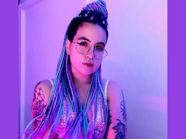 SammyBeil with blue and purple braids, glasses, nose ring, and tattoos, wearing a white top, has a serious expression, purple and pink lighting, and a topknot hairstyle.
