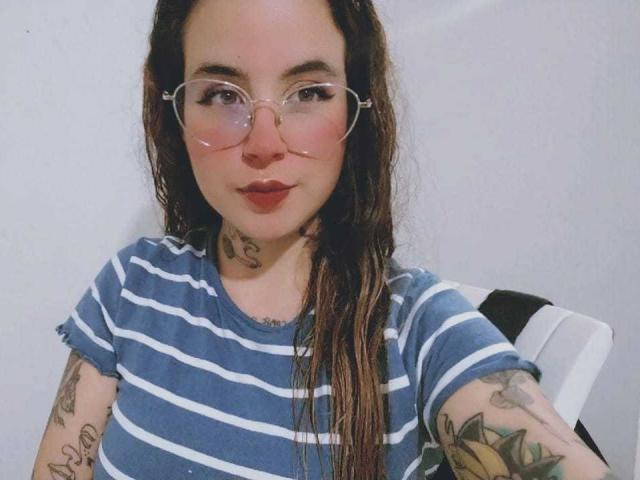 SammyBeil with wet brown hair, wearing clear glasses and a blue-striped shirt, showing tattoos on her neck and arms, with a subtle pout and red lipstick.