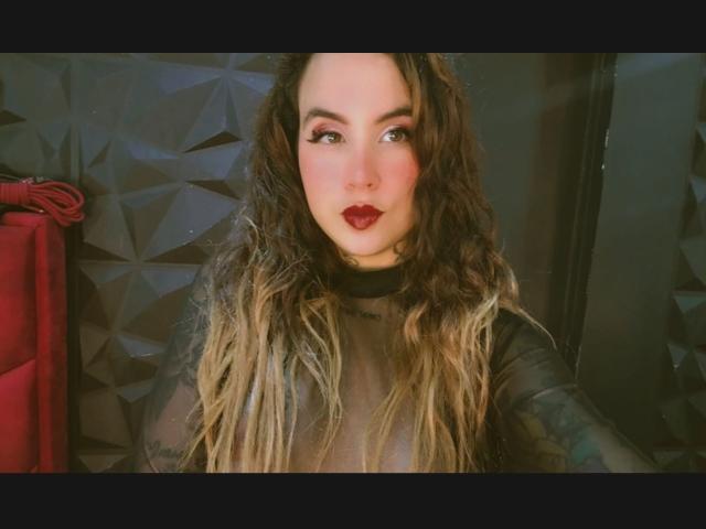 SammyBeil has long, wavy, blonde-tipped brown hair, red lipstick, and is wearing a sheer black top, looking at the camera with a slightly pouty expression.