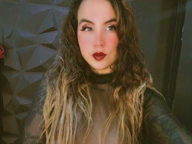 SammyBeil with wavy brown and blonde hair, wearing a sheer black top, has red lipstick and dark eye makeup, looking at the camera with a slight smile.
