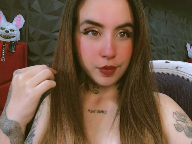 SammyBeil with long brown hair, wearing no clothes, showing tattoos on her chest and arm, holding hair, with red lipstick, and a cute stuffed cat in the background.