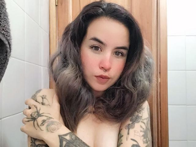 SammyBeil with shoulder tattoos, nose ring, and gray wavy hair, covering her tits with one hand, in a bathroom. Her ass is not visible.