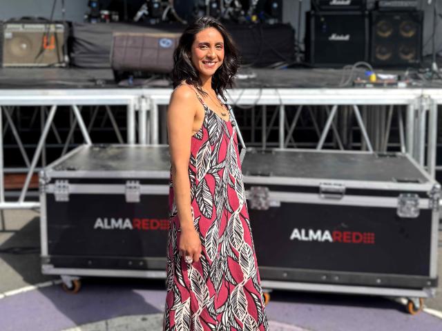 MiaAmoree stands in the foreground, smiling. She's wearing a sleeveless, patterned dress with pink, white, and black abstract designs. She has wavy black hair and is positioned slightly to the right. The background features two black AlmaRedDiB metal cases with white and red logos, and a metal stage with equipment. The image is brightly lit with natural light. The composition focuses on MiaAmoree and the patterned dress. The overall style is casual and contemporary.